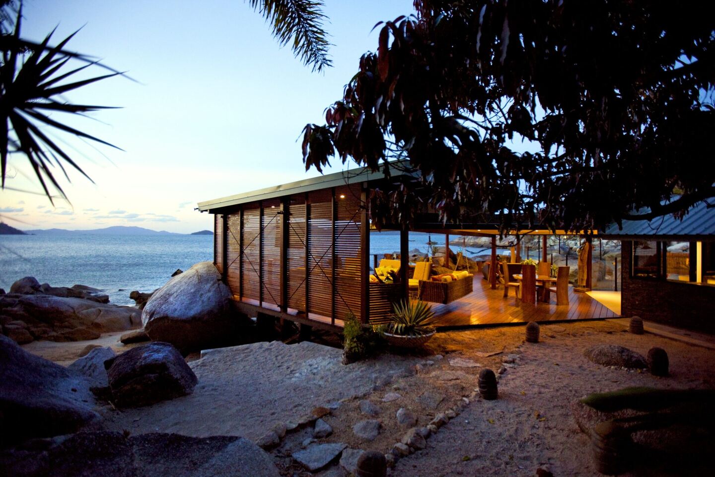 The house nestled into rainforest above the beach, seen from the water at dusk
